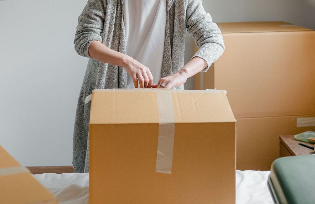 Slim female in cardigan unpacking cardboard box put on bed close to suitcase while standing near window in attic style room in daylight