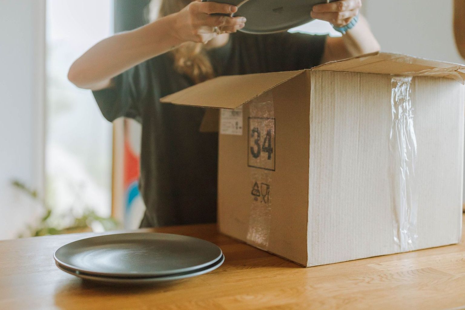 A woman unpacks ceramic plates from a cardboard box on a wooden table in a cozy indoor setting.