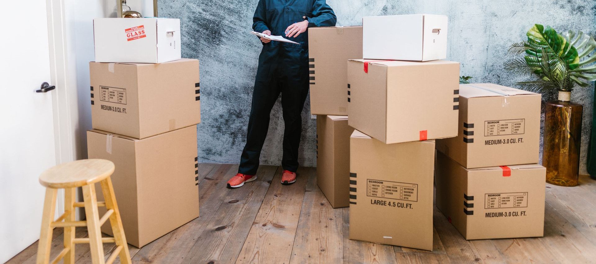 Smiling mover in coveralls standing with parcels, clipboard in hand, indoors.