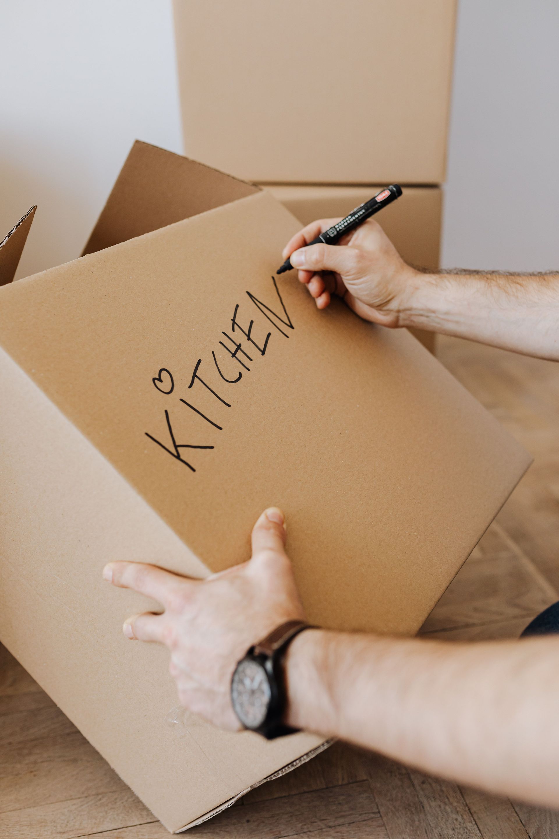 Crop unrecognizable male using black marker to write on carton container word kitchen and draw heart to identify box with packed kitchenware during relocation